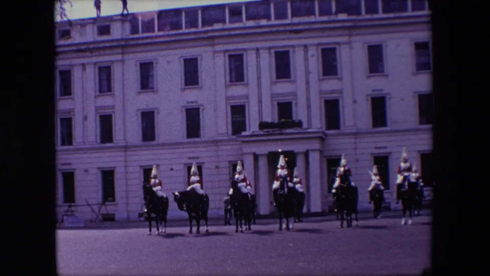 A wide visitor-shot frame shows mounted guards spread across a broad paved courtyard in front of a multi-story pale building, with small figures visible along the roofline or upper edge.