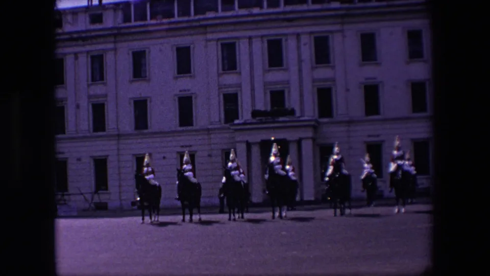 1967 London 8mm frame showing a darker view of mounted ceremonial soldiers across a courtyard, with the stone building and windows behind them.