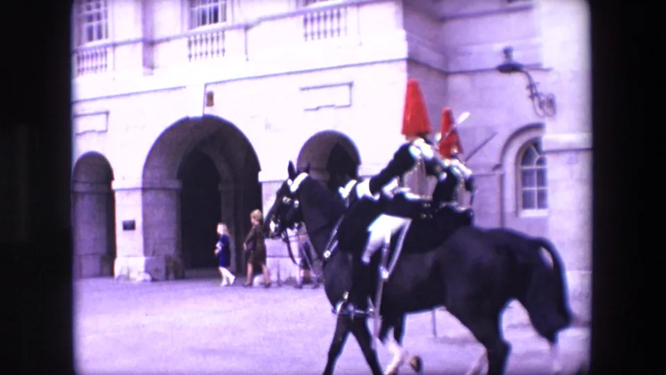1967 London frame showing close mounted cavalry riders in ceremonial uniform with red plumes and swords moving past arched stone entrances as pedestrians stand nearby.