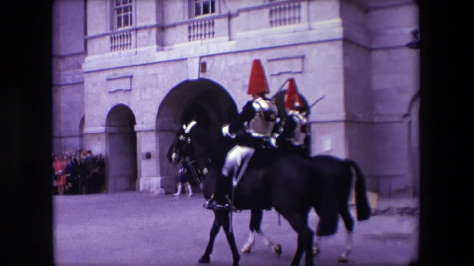 In 1967 London home-movie footage, two ceremonial riders in reflective armor and red plumed headdresses ride dark horses past a formal stone building as spectators watch nearby.