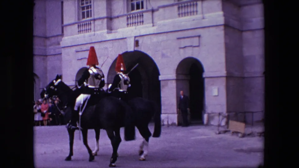 Two riders in ceremonial armor and red plumes move on horseback outside a large stone building in London, with a small crowd watching from the edge of the entrance area.