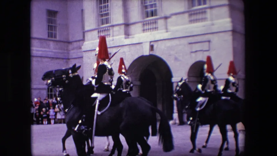 Mounted riders in ceremonial armor and red plumes pass through the frame outside a formal London stone building, with spectators gathered near the arched entrance in the background.
