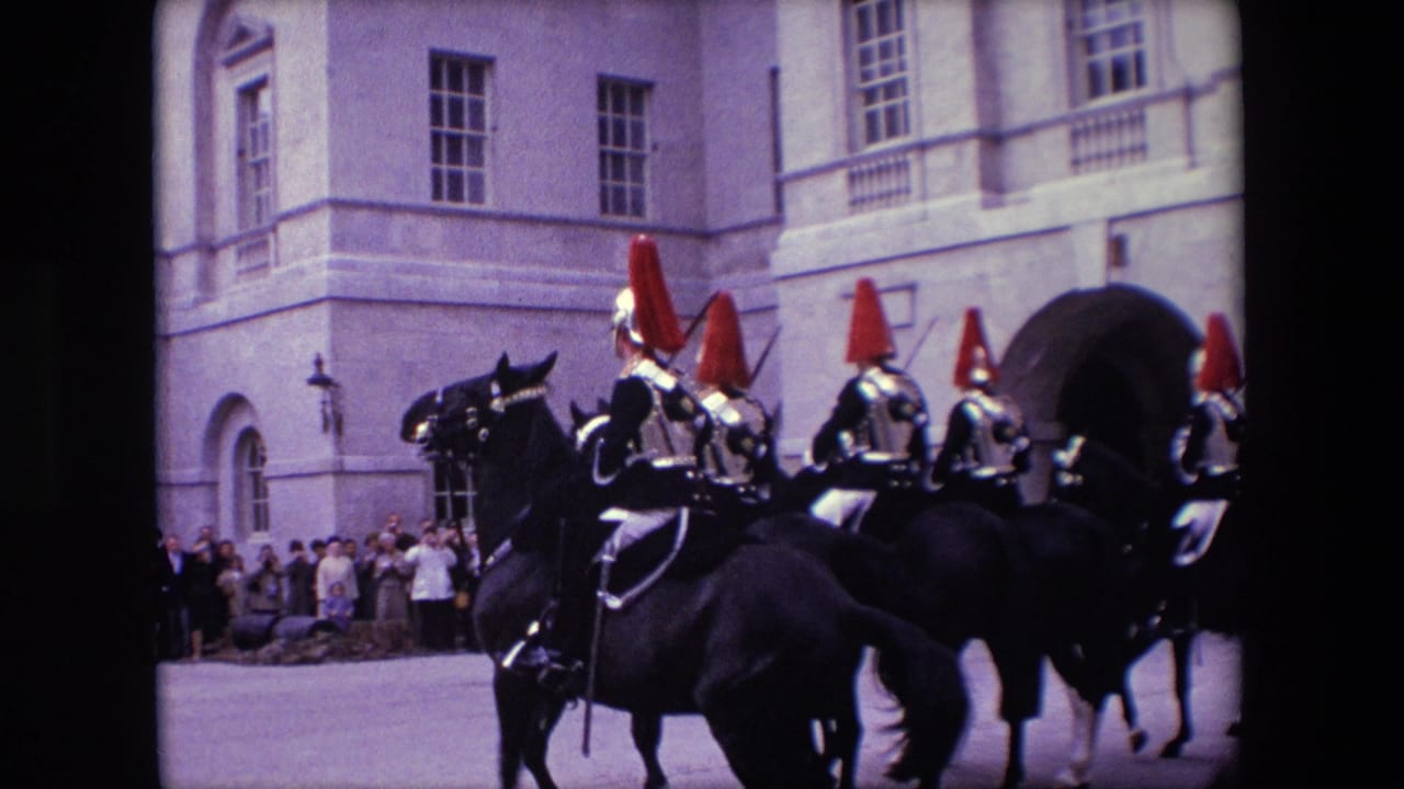 1967 London home-movie frame showing mounted soldiers in ceremonial uniforms riding dark horses past spectators near a pale stone building with arched openings.