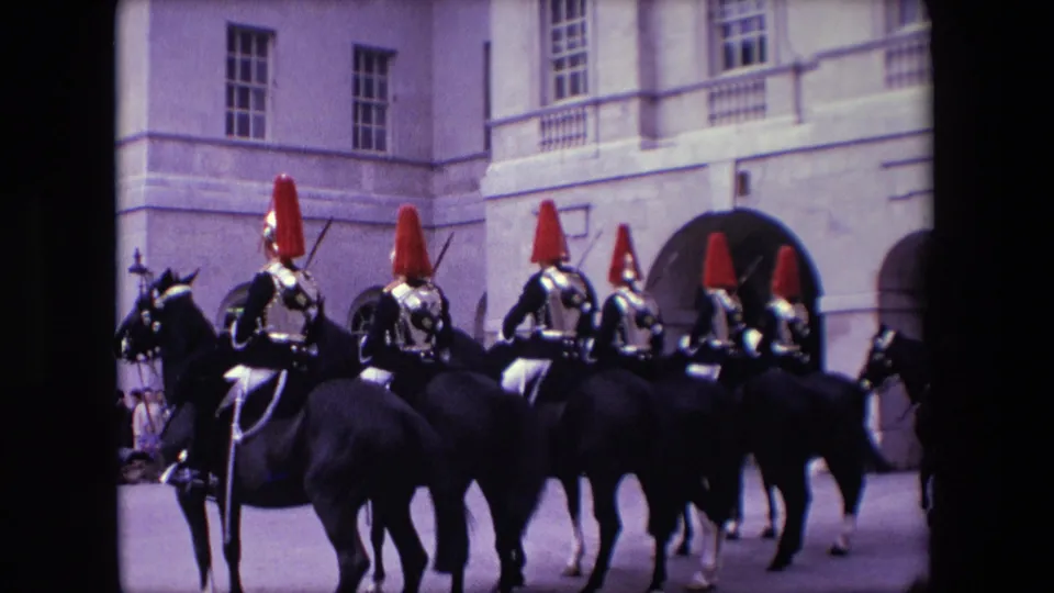 1967 London home-movie still showing a line of ceremonial mounted soldiers on dark horses passing a pale stone building while a crowd stands in the background.