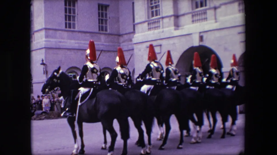 1967 London frame showing mounted soldiers in ceremonial uniform riding in a line before a large stone building with arches, windows, and spectators along the edge.