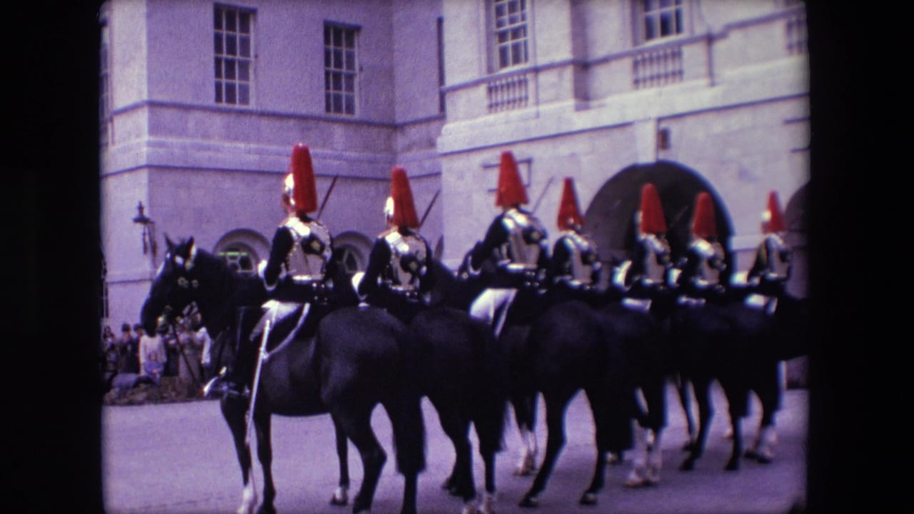 Mounted ceremonial riders with red plumed helmets pass a stone building as spectators watch.