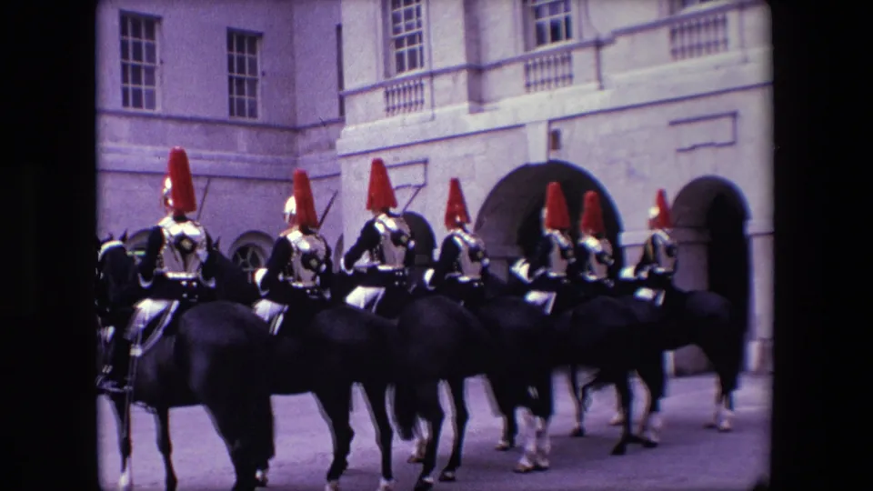 A handheld 8mm view shows ceremonial riders on dark horses moving past a stone building in London, with red helmet plumes and reflective armor catching the light.