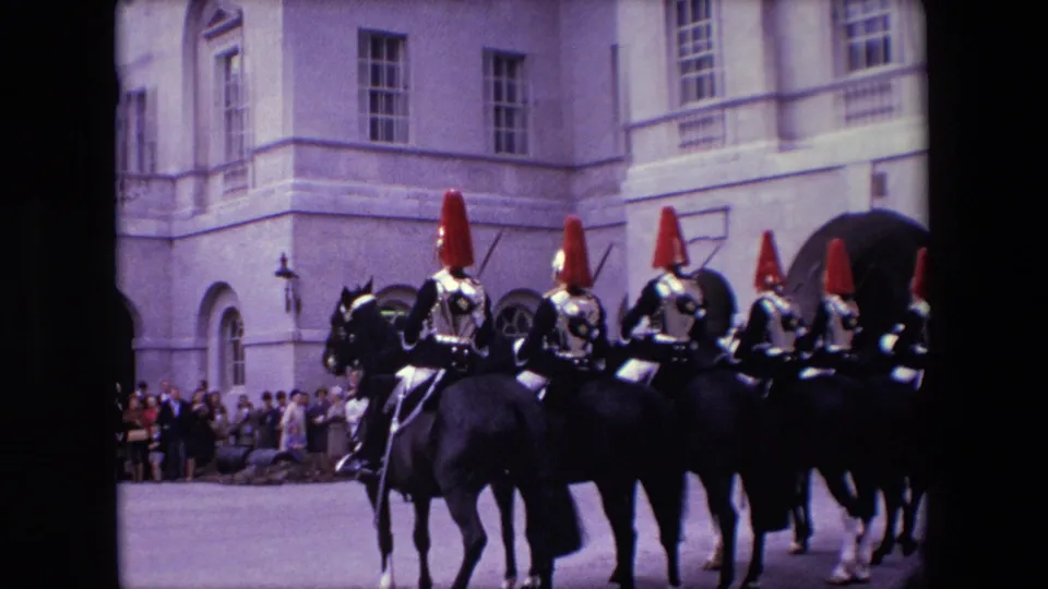 1967 London home-movie frame with mounted uniformed guards riding in formation near spectators gathered along a large stone facade.