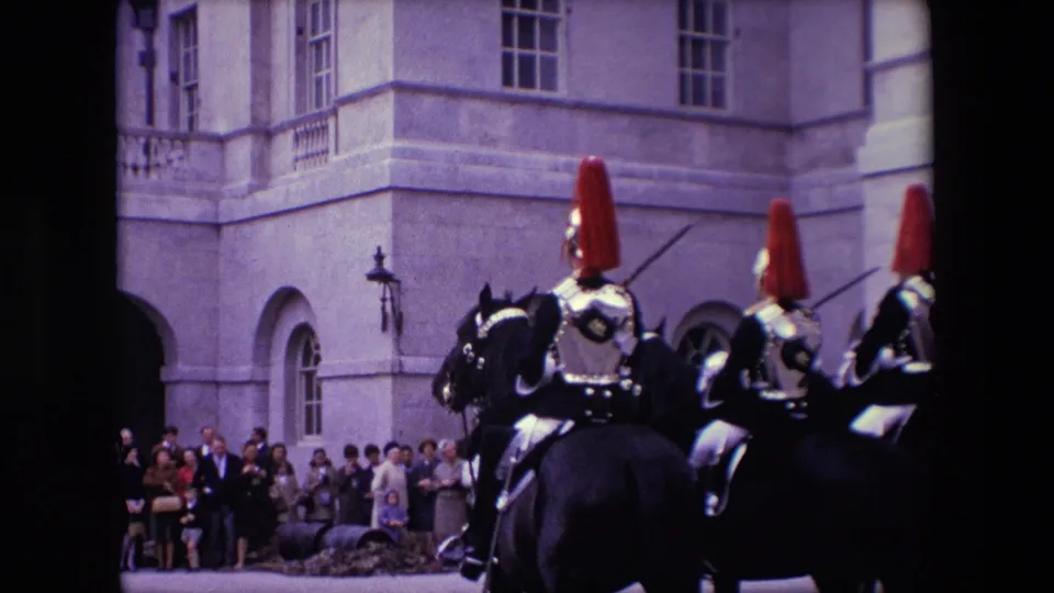 1967 London home-movie frame showing spectators beside a stone building while mounted guards and horses pass close to the camera.