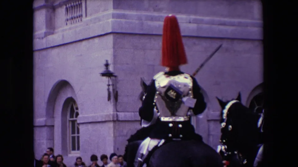 1967 London home-movie frame showing a close rear view of a mounted uniformed guard with a red plume, with other horses and spectators near a stone building.