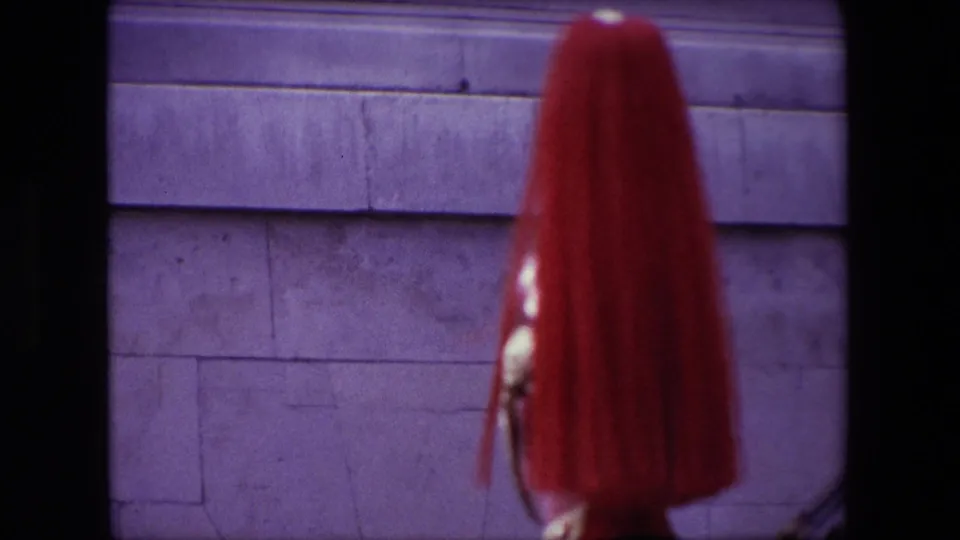 1967 London 8mm frame with a close, soft-focus view of a ceremonial guard's red plumed helmet and armor edge against a stone wall.