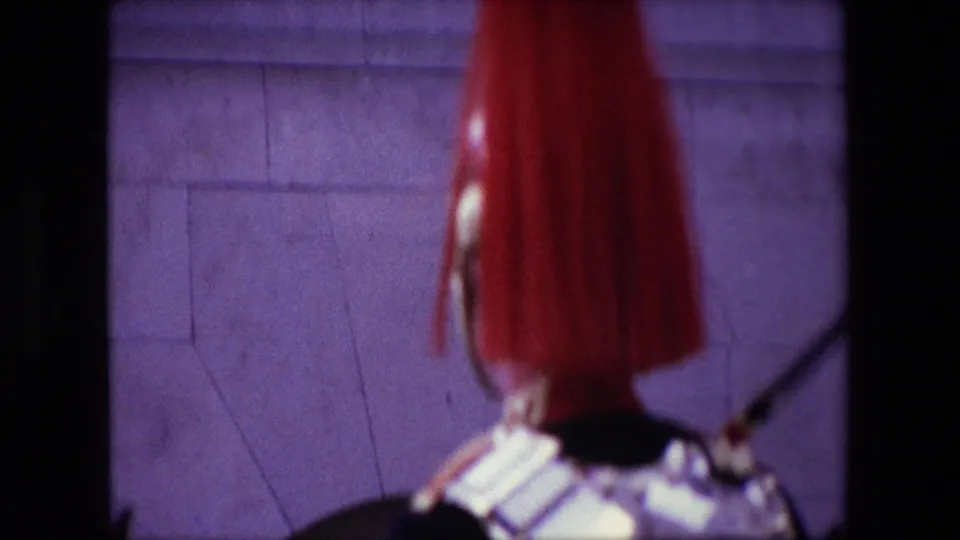 1967 London home-movie frame with a blurred ceremonial guard in the foreground and a stone building wall behind, suggesting the camera is positioned near the passing formation.