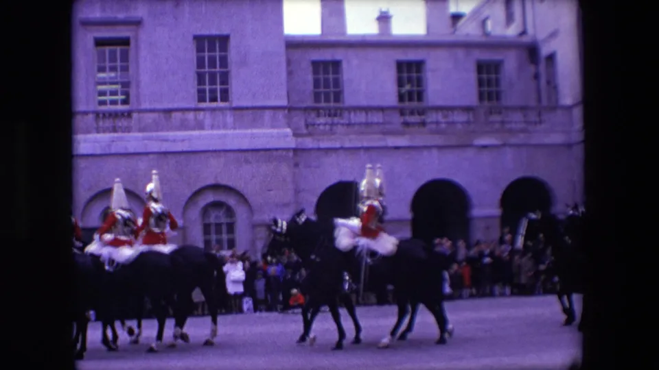 Mounted ceremonial guards and dark horses continue past a crowd-lined stone facade in the final frame of the 1967 London home-movie clip.
