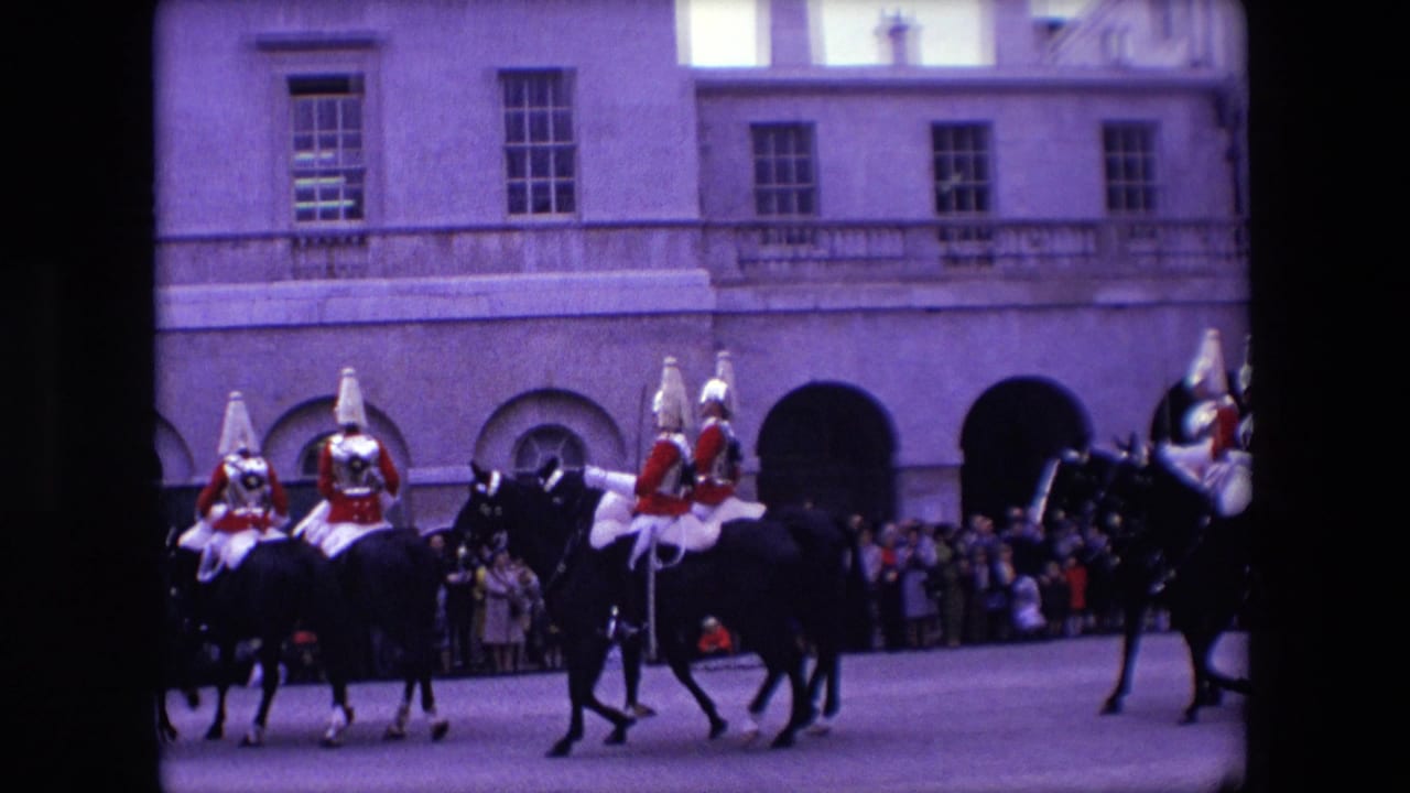 In 1967 London, mounted guards in red ceremonial uniforms ride dark horses past a watching crowd beside a pale stone building with arched openings.