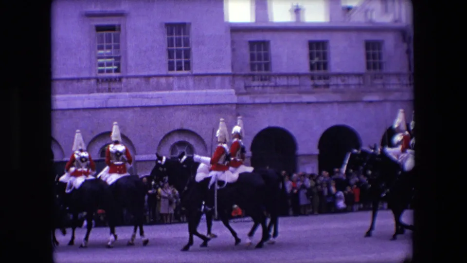 In 1967 London, mounted guards in red ceremonial uniforms ride dark horses past a watching crowd beside a pale stone building with arched openings.
