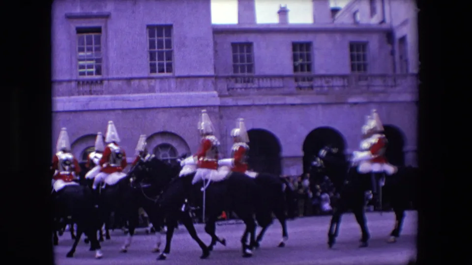 Several mounted guards in ceremonial dress ride through a London courtyard as spectators watch from the edge of the route near a formal stone facade.