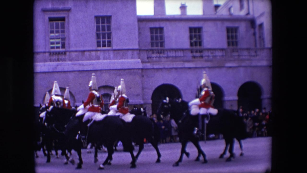Mounted soldiers in dress uniform ride past a crowd in front of an arched stone building.