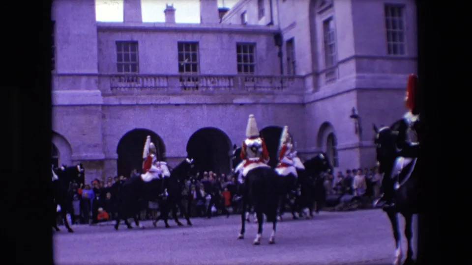 1967 London frame with mounted ceremonial cavalry near the edge of the shot, spectators along the street, and a historic building behind them.