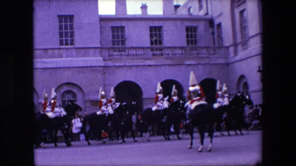 1967 London 8mm frame showing mounted uniformed riders passing in front of spectators, with pale stone walls and arched entrances behind them.