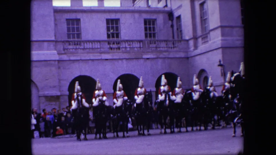 Mounted guards in ceremonial dress line the foreground outside Buckingham Palace, with visitors gathered along the edge of the court.