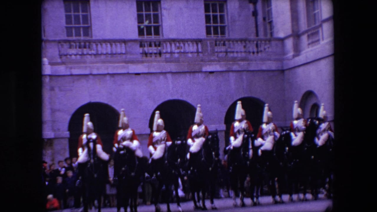 1967 London home-movie frame showing British mounted palace guards in red ceremonial uniforms moving in formation before arched stone buildings.
