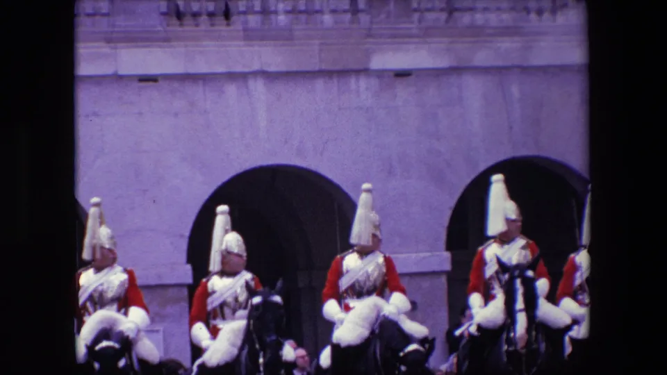1967 London frame with mounted palace guards in ceremonial red uniforms and plumed helmets passing in front of large arched openings.