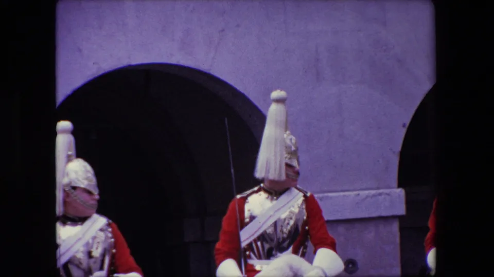 Close 1967 London home-movie view of ceremonial mounted guards, with red sleeves, pale plumes, metal helmets, and a palace arch behind them.