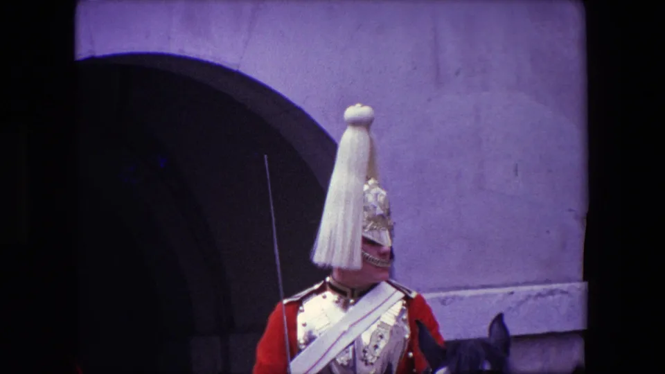 1967 London frame centered on a mounted palace guard wearing a plumed ceremonial helmet and red uniform in front of a large stone arch.