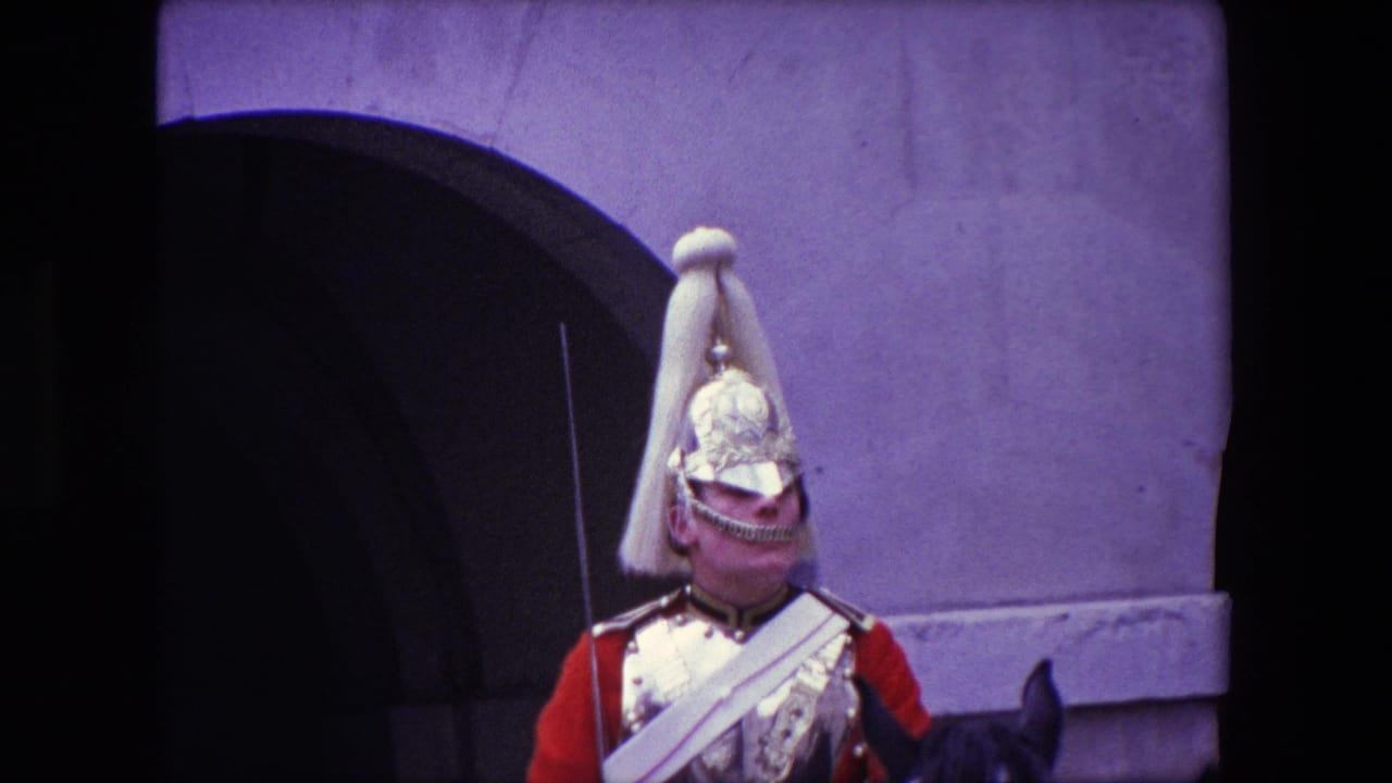 1967 London home-movie frame showing a mounted ceremonial guard in a red tunic and polished helmet near a stone archway.