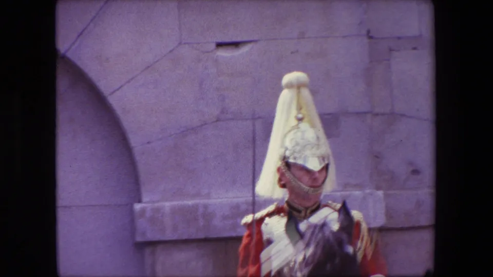 1967 London home-movie frame showing a mounted ceremonial guard in red and silver uniform passing a pale stone wall and arch detail.