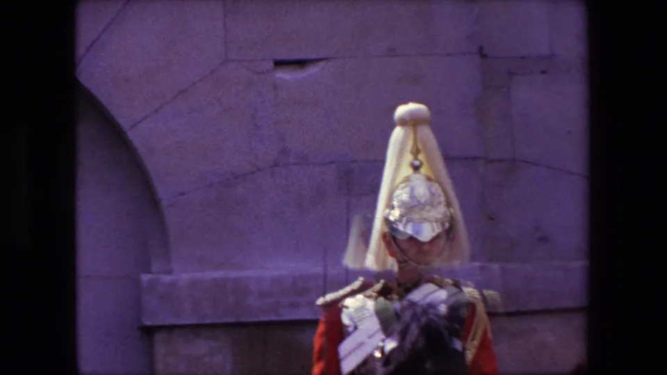 A London ceremonial guard on horseback faces the camera, with a reflective helmet, pale plume, red sleeve, and shoulder decoration visible in the 1967 8mm frame.