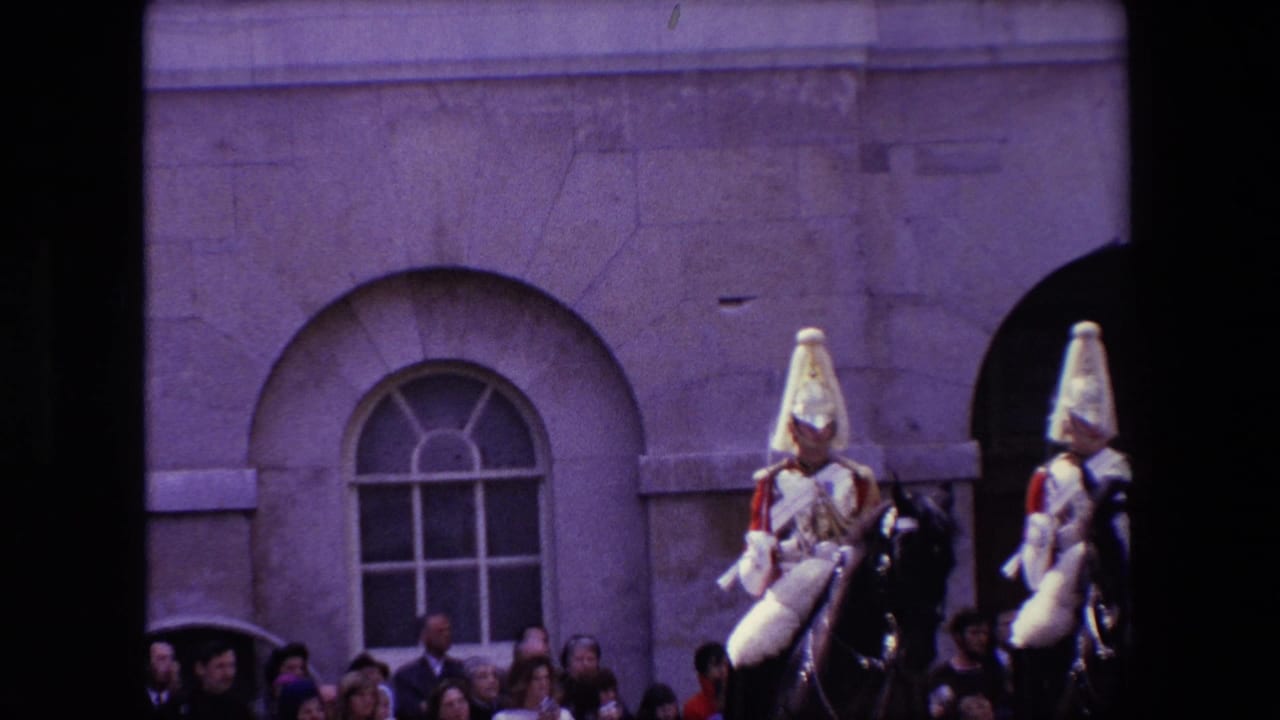 Two mounted guards ride before spectators along a stone building.