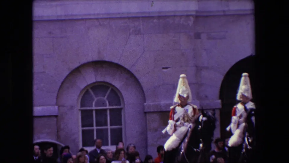 Two mounted ceremonial guards in plumed helmets ride past a watching crowd beside a stone London building, with arches and windows visible in the background.