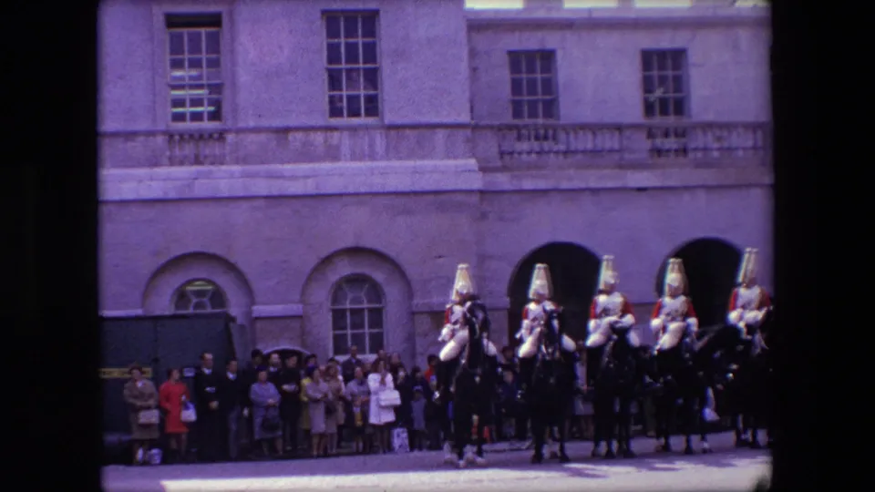 Several mounted ceremonial guards in London hold swords while riding in formation before spectators along a stone building facade in 1967 home-movie footage.