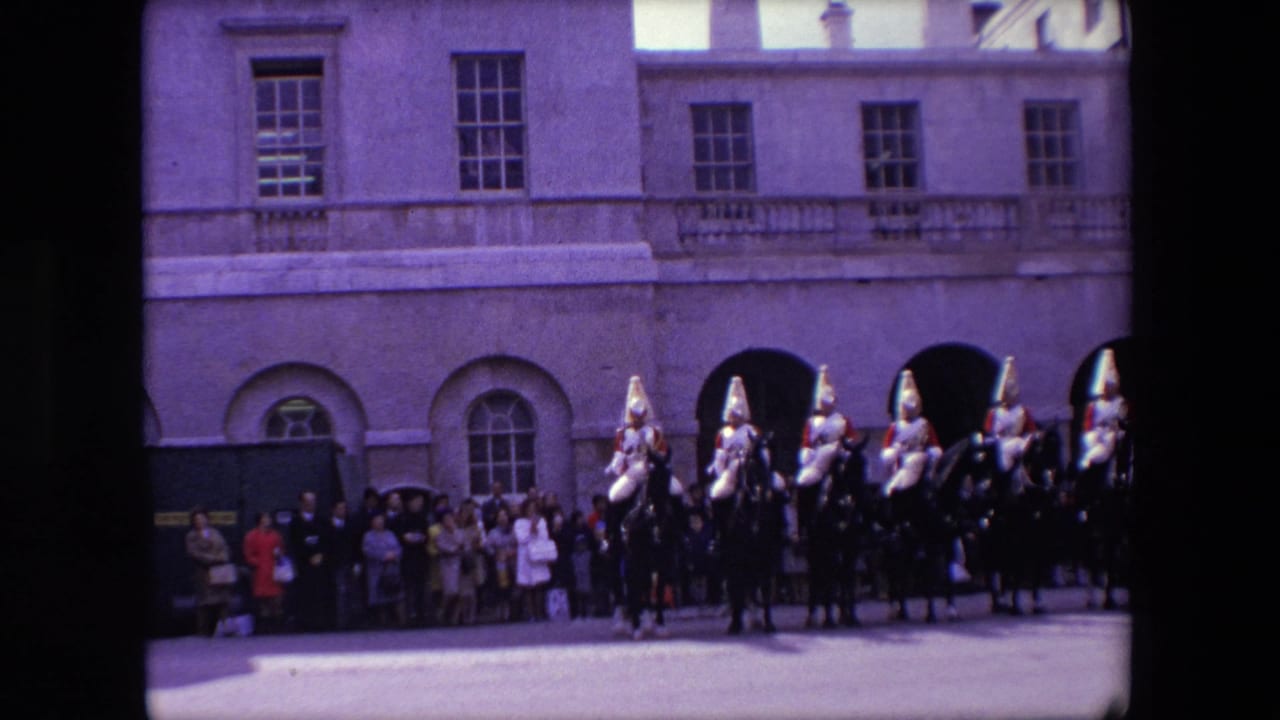 1967 London home-movie frame showing mounted ceremonial guards on dark horses with spectators lined up against a formal stone facade.