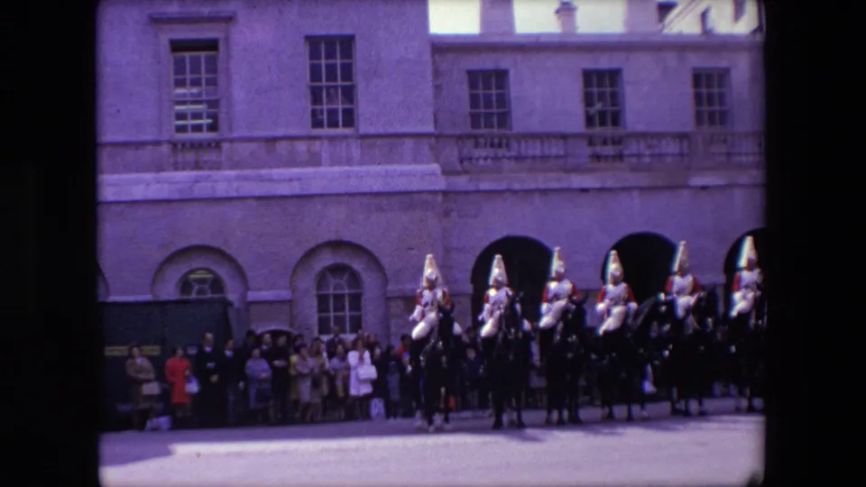 1967 London home-movie frame showing mounted ceremonial guards on dark horses with spectators lined up against a formal stone facade.