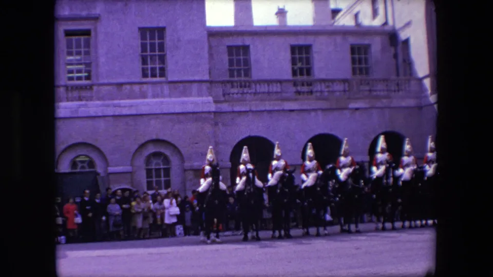 1967 London frame with a line of mounted ceremonial guards near arched openings while a small crowd watches from the building edge.