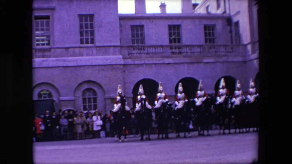 1967 London home-movie view of mounted guards in ceremonial helmets beside spectators at the base of a large stone building.