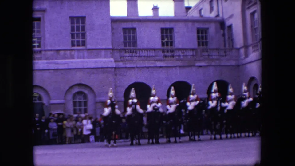 1967 London 8mm frame showing mounted ceremonial riders in plumed helmets moving past spectators outside a formal stone building.