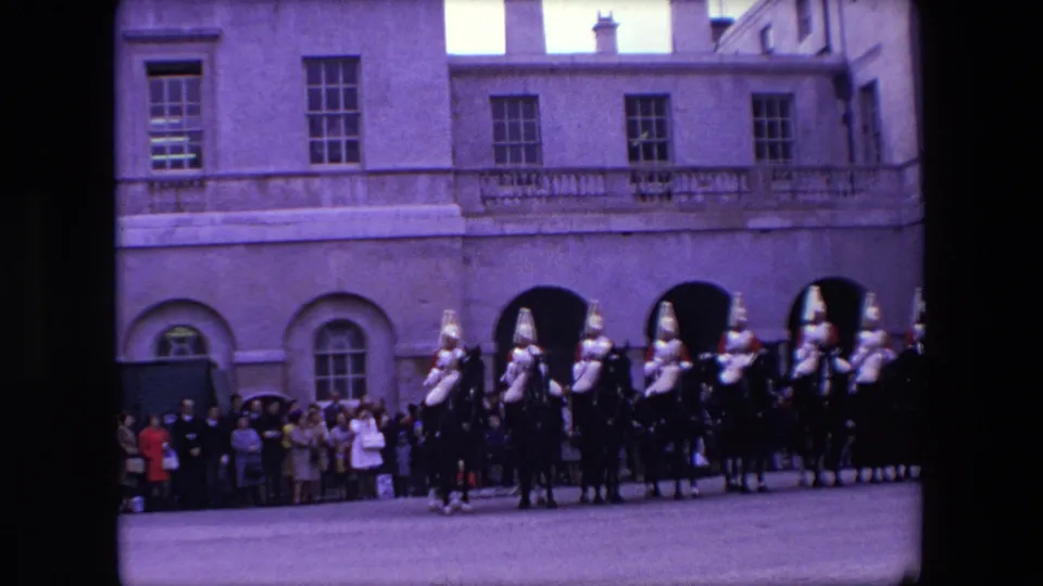 1967 London 8mm frame with ceremonial riders on horseback passing a dense line of spectators near arched doors and tall windows.