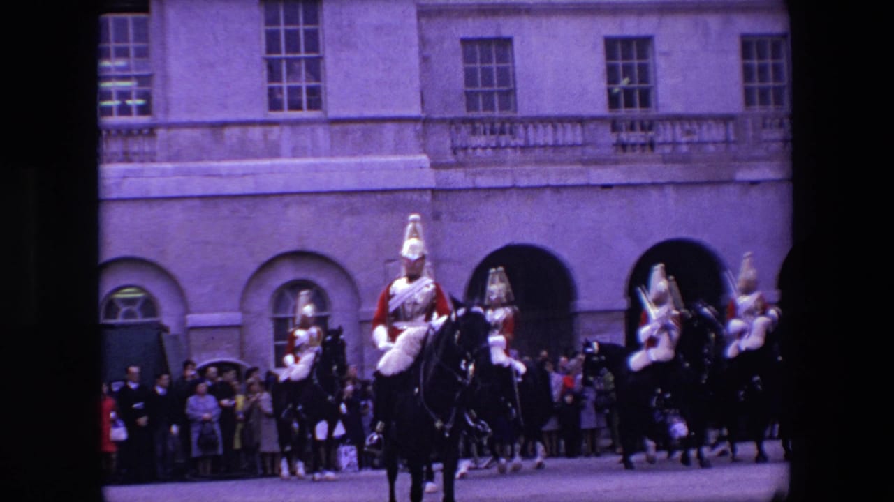 1967 London home-movie frame showing mounted Queen's Guard soldiers in red dress uniforms riding past spectators beside a pale stone building.
