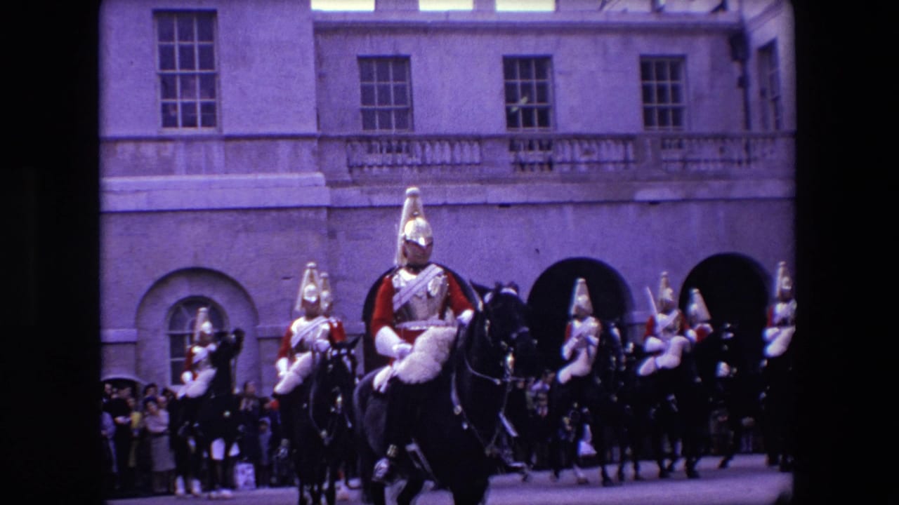 Mounted guards in red uniforms ride in formation before a crowd and stone building.