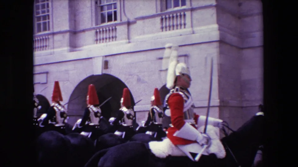 Closer 8mm frame of a mounted guard in red uniform and white plume passing other helmeted riders near a large arched entrance in London.