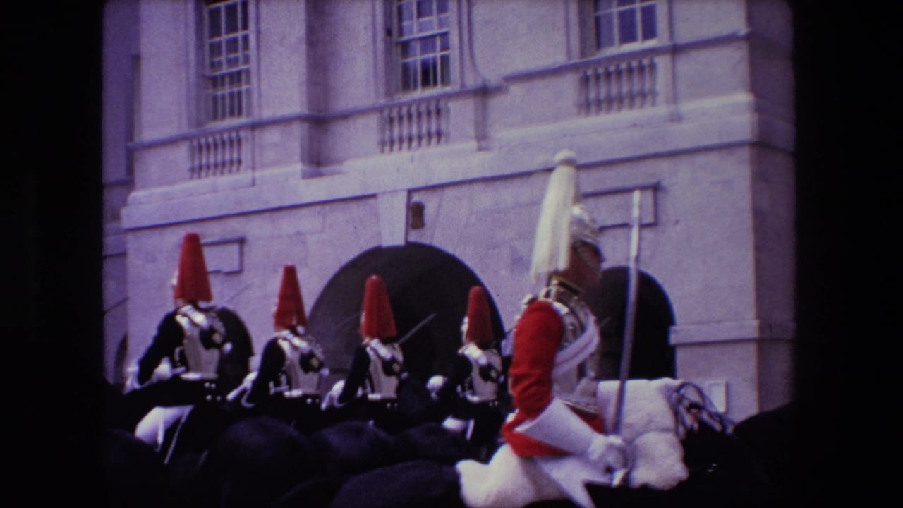 1967 London home-movie frame showing mounted ceremonial guards in red uniforms and plumed helmets moving past a pale stone facade with arched entrances.