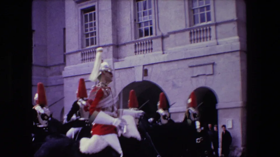 1967 London home-movie frame with a mounted guard in red and white ceremonial dress in the foreground and other helmeted riders behind him near arched stone entrances.