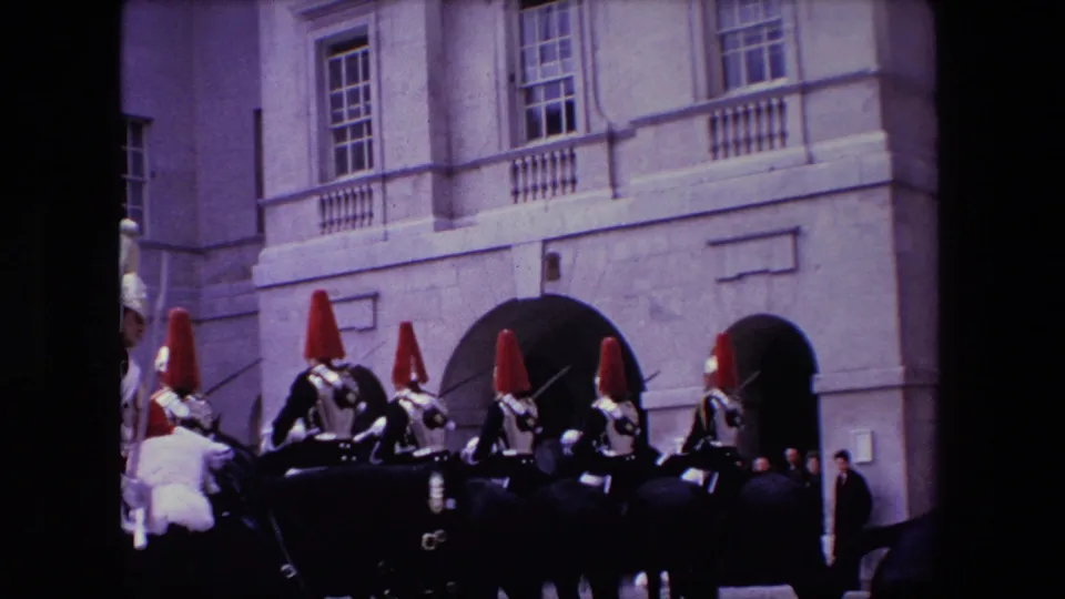 1967 London home-movie frame showing a row of mounted ceremonial guards in plumed helmets before a pale stone building with upper windows and arched openings.