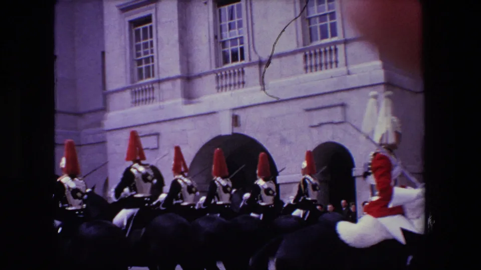 1967 London home-movie frame showing mounted ceremonial guards in formation with a red-uniformed rider partly cropped at the right edge and a stone building behind them.