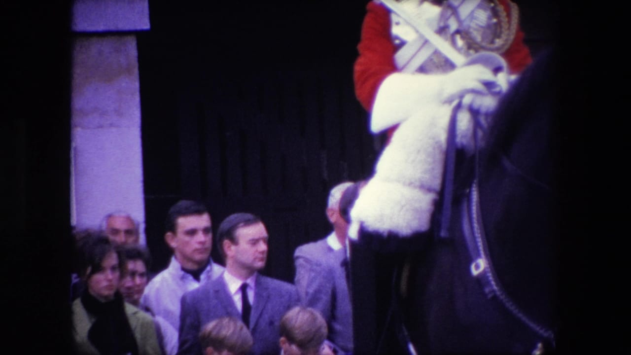 1967 London home-movie frame with adult spectators and children watching near a mounted ceremonial rider, cropped by the edge of the horse and uniform.