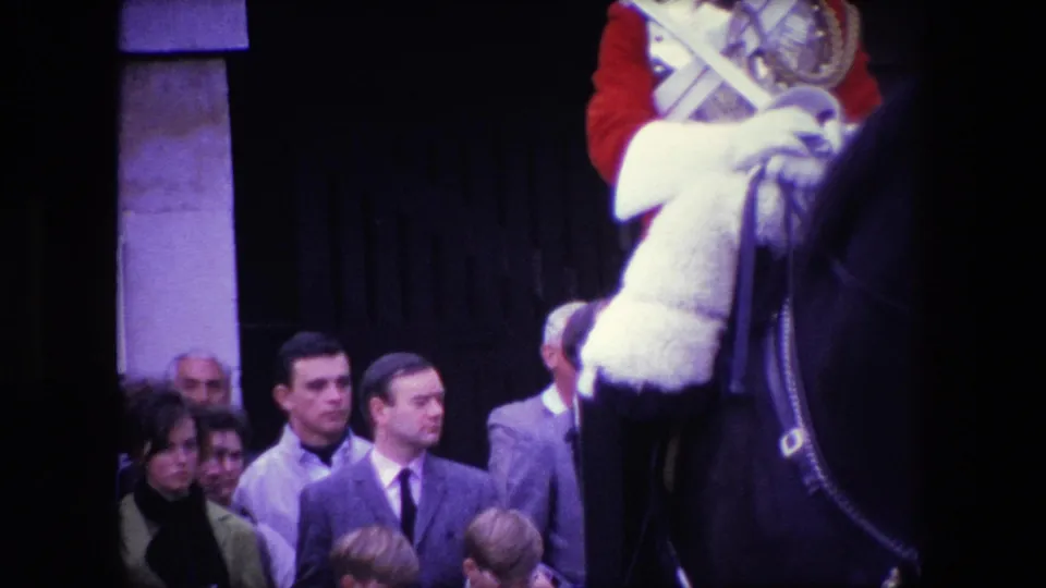 1967 London home-movie frame with adult spectators and children watching near a mounted ceremonial rider, cropped by the edge of the horse and uniform.