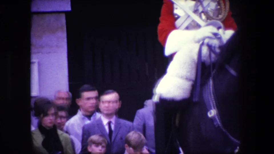 1967 London home-movie frame showing the horse and cropped uniformed rider close to a crowd of adults and children near a stone entrance.